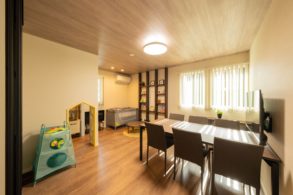 明るいダイニングとプレイルームが一体となった部屋。長いテーブルには椅子が並び、壁際には本棚が設置されています。左側には小さな子ども用のプレイエリアがあり、おもちゃやベビーベッドが配置されています。大きな窓から光が差し込み、温かみのある空間です。