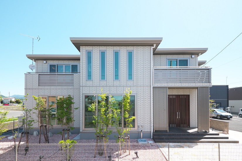 大館展示場、パルフェNの外観写真。白い磁器タイル外壁が特徴的な二階建ての住宅。縦長の窓とバルコニーがあり、前庭には若い木々が植えられています。明るい青空が背景に広がっています。