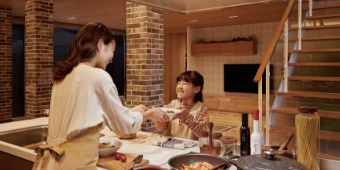 宿泊体感棟紹介ページへのリンク画像です。広々としたキッチンで母親が料理を渡し、娘が笑顔で受け取っている様子。暖かい照明と木目調のインテリアが落ち着いた雰囲気をつくっています