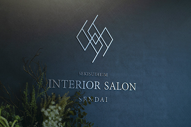 受付イメージ画像。SEKISUIHEIM INTERIOR SALON SENDAIと書かれた壁のサイン。暗めの背景にシンプルでスタイリッシュなデザインが施されており、左下に植物が飾られています。