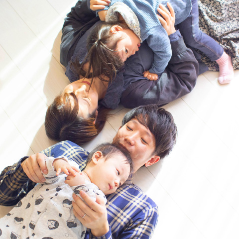家の中で、床に寝転んで寄り添う家族の写真。お父さんとお母さんが赤ちゃんを優しく抱きしめ、上に小さな子どもが重なるようにして微笑んでいます。