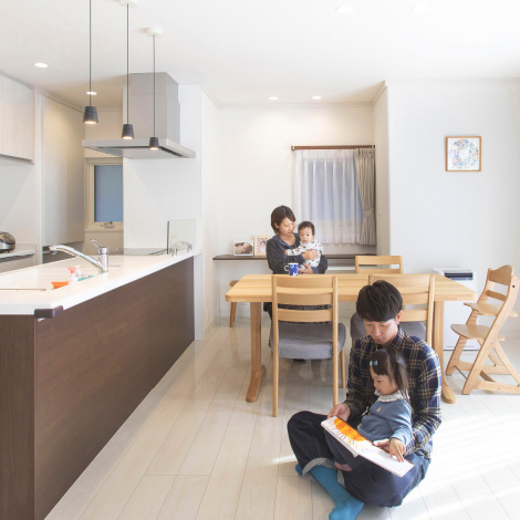 after followイメージ画像。明るいリビングダイニングで家族がくつろいでいます。父親が床に座り、子どもに絵本を読んでいる一方、母親はもう一人の子どもを抱きながら食卓に座っています。