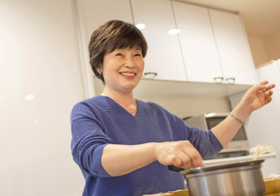 eventuallyイメージ画像。女性がキッチンで鍋を持ちながら、笑顔で料理をしています。明るい雰囲気の中で、楽しそうな表情が印象的です。