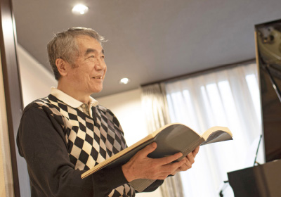 piano spaceイメージ画像。男性が楽譜を手に持ち、微笑みながら読んでいる様子。後ろにはピアノがあり、音楽を楽しんでいる雰囲気です。