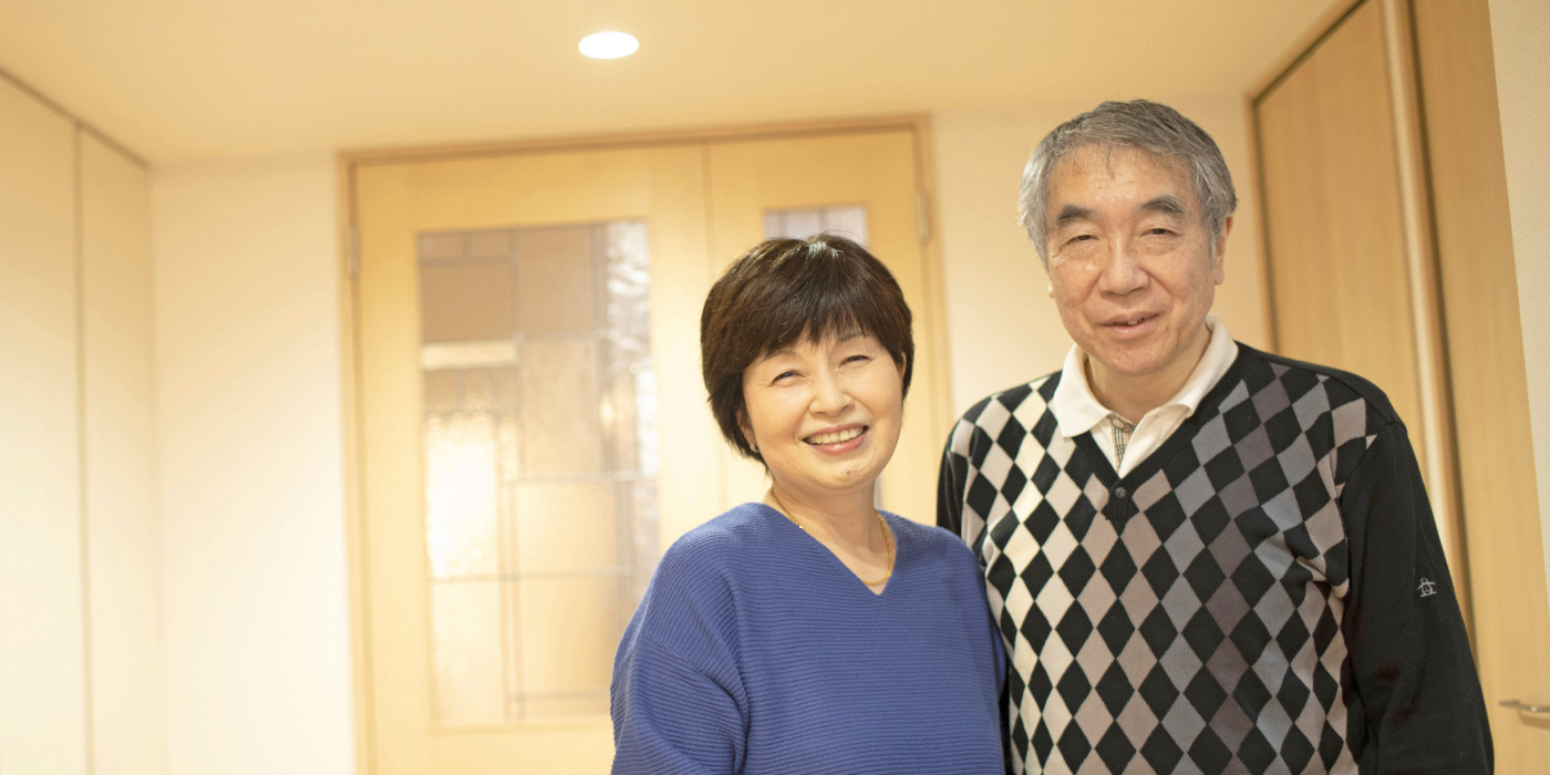 インタビュー02イメージ画像。年配の男女が室内で笑顔を見せています。女性は青いセーター、男性はダイヤ柄のセーターを着ており、暖かく穏やかな雰囲気が漂っています。