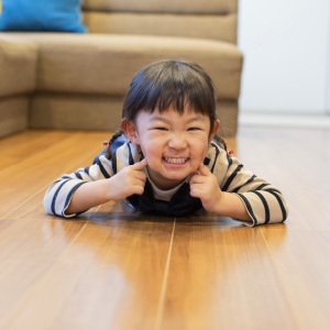 フロアプランコレクションイメージ画像。床にうつ伏せになって、楽しそうに笑っている幼い女の子です。両手で頬を押さえ、元気いっぱいの笑顔を見せています。背後にはソファが見え、明るいリビングで遊んでいる様子が伝わる一枚です。