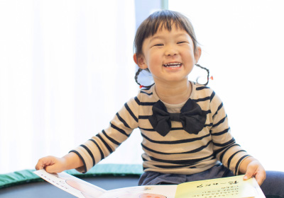 Good timingのイメージ画像。ストライプの服を着た女の子が、絵本を広げてにこやかに笑っています。小さな手で本をめくりながら、楽しそうな表情がとても印象的です。明るい光が差し込む部屋で、読書を楽しむ微笑ましい姿です。