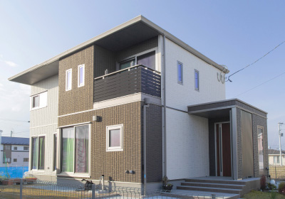 Reliefのイメージ画像。茶色と白の外壁が美しい二階建ての住宅。広いバルコニーや窓があり、玄関に続く階段が家のデザインを引き立てています。青空の下で、静かな住宅街に建つ印象的な家です。