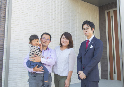 イメージ画像。新しい家の前で撮影された家族写真です。父親が娘を抱え、母親が隣に立って笑顔を見せています。スーツ姿の担当営業の男性が一緒に写っており、家族をサポートするような雰囲気です。