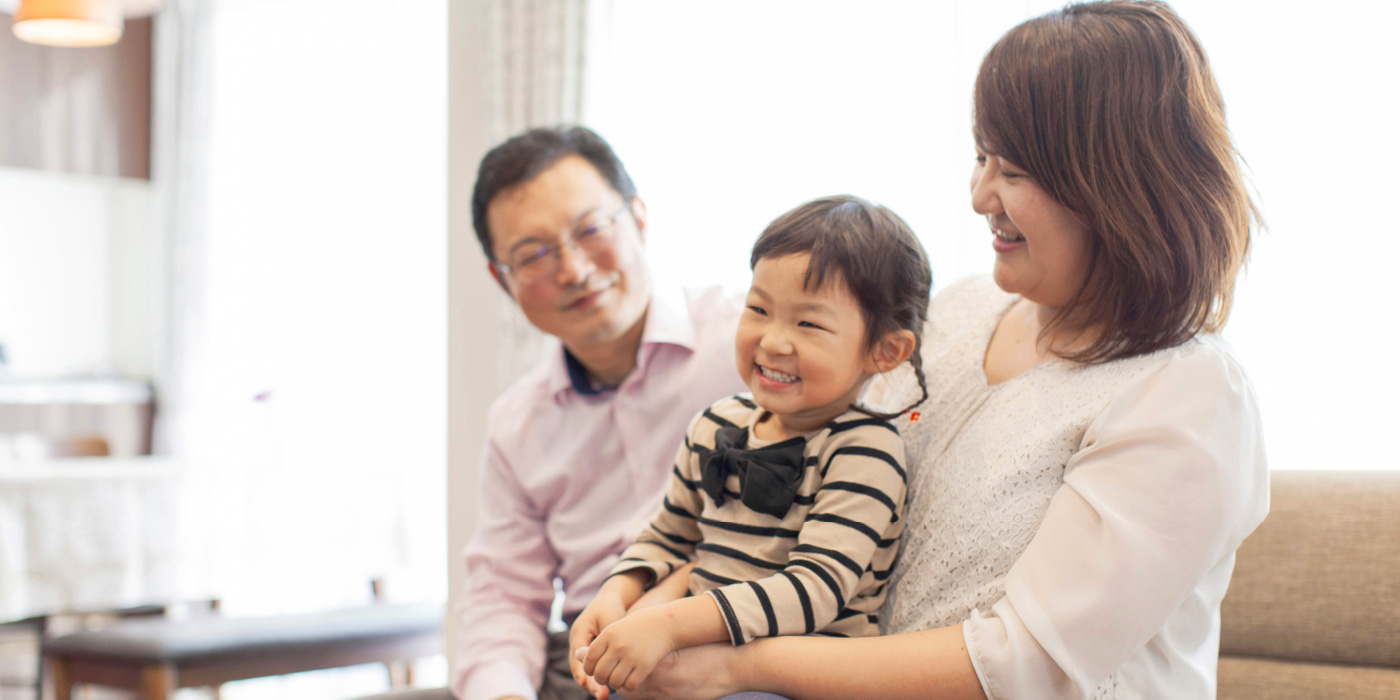 インタビュー03イメージ画像。ソファに座る家族の写真。お母さんが膝の上に小さな女の子を抱きかかえ、女の子は満面の笑みを浮かべています。お父さんは少し離れたところから優しそうに二人を見守っていて、明るい部屋の中で、家族の温かな雰囲気が伝わる一枚です。