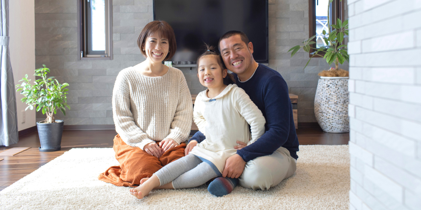 インタビュー04イメージ画像。リビングのカーペットの上に座る家族。両親が笑顔で、真ん中にいる娘を囲んでいます。背後にはテレビと観葉植物があり、温かい光が差し込む中、家族の絆が感じられるリラックスした一枚です。