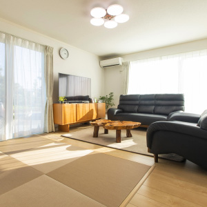 フロアプランコレクションイメージ画像。広々としたリビングには、大きな窓から光が差し込んでいます。黒いソファと木製のテレビ台、特徴的な木製テーブルが配置され、部屋全体が明るく落ち着いた雰囲気で、畳の床が和の要素を加えています。