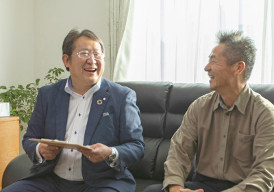 Encounterイメージ画像。‘ソファに座り、笑顔で談笑する二人の男性。スーツ姿の担当営業の男性が書類を手に持ち、施主様と和やかに会話しています。窓から光が差し込み、明るい室内での親しい交流が感じられます。