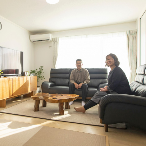 Warm houseイメージ画像。リビングのソファに座るご夫婦。中央には木製のテーブルがあり、テレビや観葉植物が置かれたリビングは柔らかな光に包まれています。落ち着いた雰囲気の中で、二人がリラックスしている様子が伝わります。