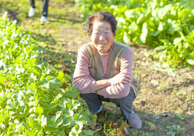 Warm houseイメージ画像。畑でしゃがんで笑顔を見せるお母様。緑の野菜が元気に育つ中、日差しを浴びて楽しそうに微笑んでいます。自然の中でリラックスした雰囲気が伝わる一枚です。