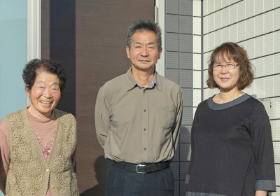 イメージ画像。家の前で並んで立つ3人の家族。左の女性は笑顔で、真ん中の男性と右の女性は穏やかな表情でカメラを見つめています。明るい日差しの中、家族の温かさが伝わる一です。