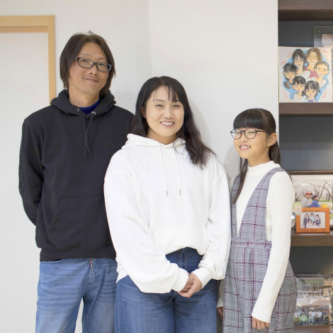 Flat loofイメージ画像。家の中で並んで微笑む3人家族。父親、母親、娘がカジュアルな服装で、リラックスした雰囲気で立っています。背景には家族の写真が飾られた棚があり、家庭的な温かさが伝わります。