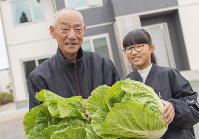 イメージ画像。老人と若い女の子が一緒に大きな白菜を持って微笑んでいて、二人は外に立ち、背後にはマイホームが見えます。収穫を喜び合うような温かい雰囲気が感じられる一枚です。
