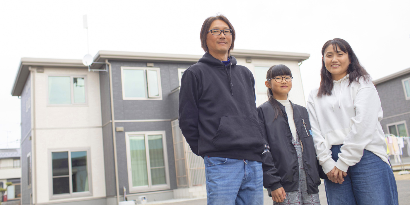 インタビュー06イメージ画像。家の前で並んで立つ家族。全員カジュアルな服装で、父親、母親、娘が笑顔を見せています。背後には二階建てのマイホームがあり、穏やかな日常の一コマが感じられます。