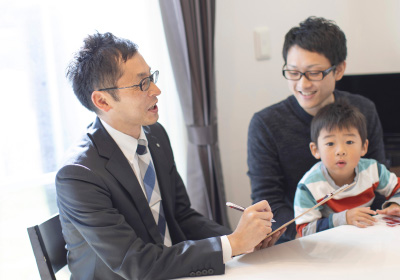 Confidenceイメージ画像。スーツを着た営業担当の男性がペンを持って説明をしている様子です。隣には父親と息子が座っており、息子は興味深そうに話を聞いています。明るいリビングで家族が話をしている場面です。