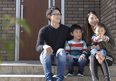 イメージ画像。家の玄関前の階段に座る家族4人。父親と母親がそれぞれ幼い息子たちと一緒に並び、楽しそうに過ごしています。小さな男の子は指を指し、家族の温かい時間が感じられる一枚です。