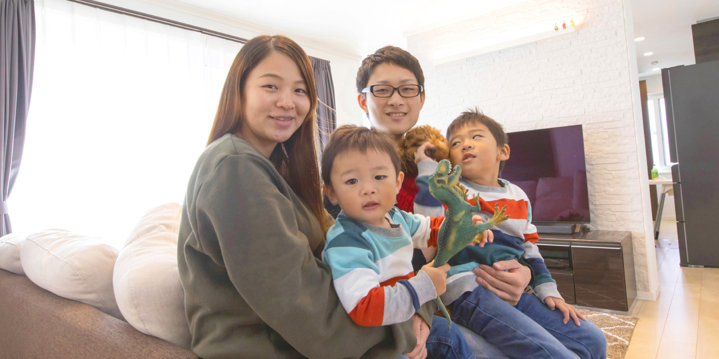 インタビュー07イメージ画像。ソファに座る家族4人が笑顔でカメラを見つめています。子供たちはおもちゃを手に持ち、楽しそうにしていて、背景にはテレビが見え、明るいリビングで家族の温かい時間を過ごしている様子が伝わります。