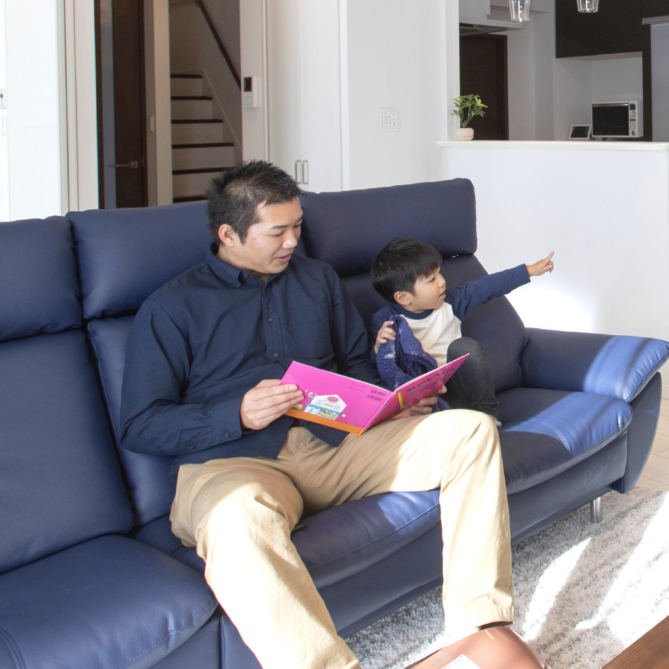 リビングルームで、お父さんがソファに座りながら子どもに絵本を読んでいる写真。子どもは指を指しながら興味津々の様子です。