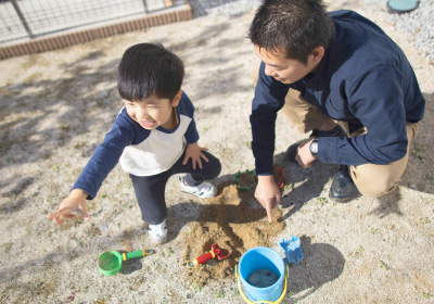 Will welcomeのイメージ写真。父親と息子が庭で一緒に砂遊びをしています。息子は楽しそうに笑いながら手を動かし、父親は砂の上に指を指して話していて、青いバケツやおもちゃが並び、晴れた日の温かい親子の時間が伝わる一枚です。
