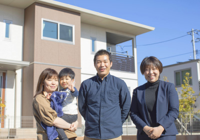 イメージ写真。家の前で笑顔の家族とスーツ姿の担当営業の女性が並んで立っている。母親が幼い息子を抱え、父親と女性は並んで立って微笑んでいて、青空の下、明るい日差しに包まれた温かい雰囲気の一枚です。