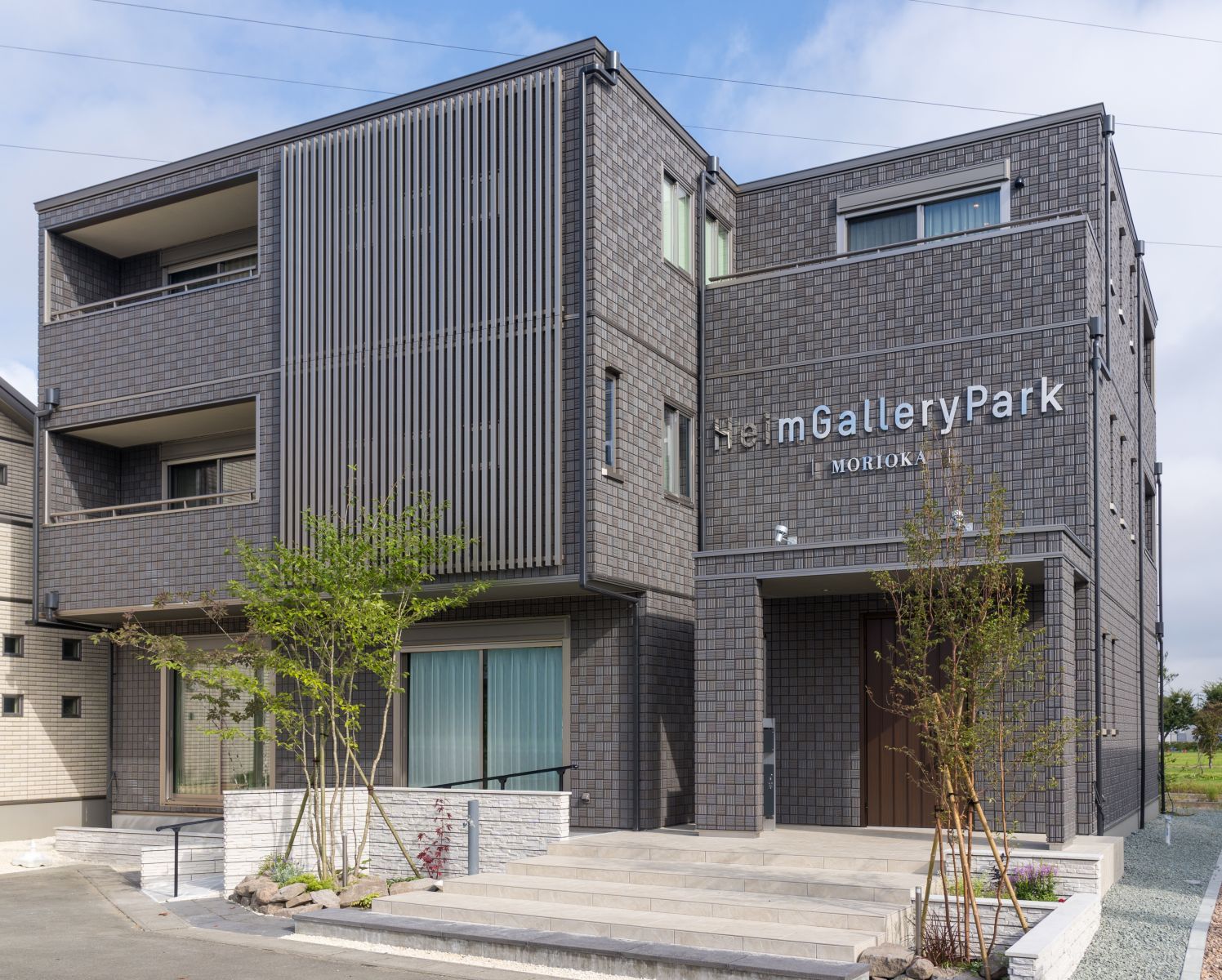 スライドイメージ画像。ハイムギャラリーパーク盛岡の外観。グレーのタイル張りの3階建ての建物で、バルコニーや大きな窓があります。建物の前には小さな木や植物が植えられており、エントランスへと続く階段も整備されています。