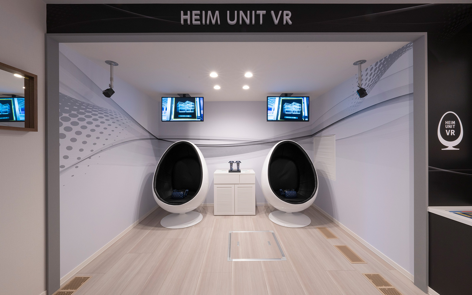 スライドイメージ画像。VR体感ゾーン。HEIM UNIT VRと書かれたスペースに、未来的なデザインの黒いクッション付き卵型の椅子が2つ並び、それぞれの前にVR機器が設置されています。上にはスクリーンが2つ吊るされています。