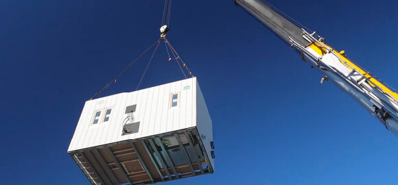 工期を大幅に短縮のイメージ画像。工事現場でクレーンを使って建物のユニットを吊り上げ、設置している様子。作業員が足場に上り、安全対策をしながら組み立て作業を進めています。空には晴れた青空が広がっています。