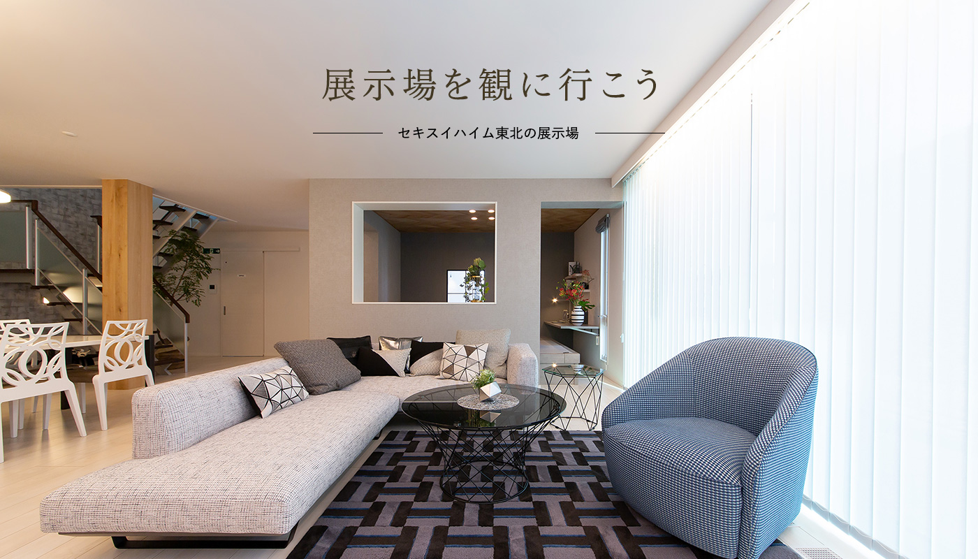 展示場情報のメインビジュアル画像です。展示場を観に行こうと書かれています。広々としたリビングルームに、L字型のソファとガラスの丸いテーブルが配置されています。窓からは明るい光が差し込み、落ち着いた空間が広がっています。