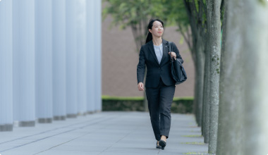 Bottomスライダー画像03スーツ姿の女性が、木々が並ぶ通りを自信を持って歩いている様子。バッグを肩にかけ、ビジネスに向かう姿が印象的です。