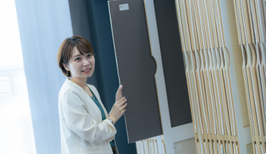 Bottomスライダー画像05展示エリアで、女性がサンプルのパネルを手に取りながらこちらを振り向いて微笑んでいます。明るい室内に整然と並んだ商品サンプルが見えます。
