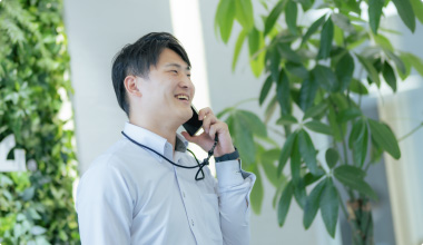 Bottomスライダー画像07若い男性が電話をしながら笑顔で話しており、背後には緑豊かな観葉植物が見えます。明るい室内で、リラックスした雰囲気です。