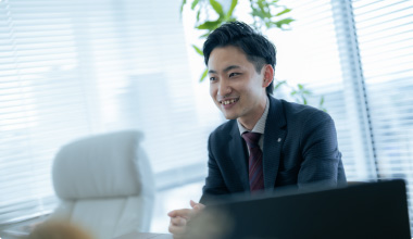 Bottomスライダー画像08スーツを着た若い男性がデスクの前で笑顔で話しています。背後には観葉植物と明るい窓があり、開放的なオフィスの雰囲気が感じられます。