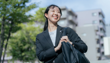 Topスライダー画像05外で笑顔の女性が、バッグを肩にかけて歩いている様子。明るい日差しの下で、背後には木々や建物が見えます。